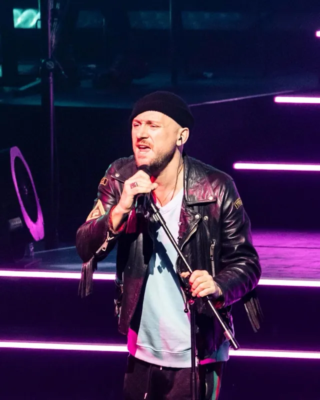 Ali Love ao vivo, de gorro e jaqueta de couro, cantando ao microfone sob luz rosa